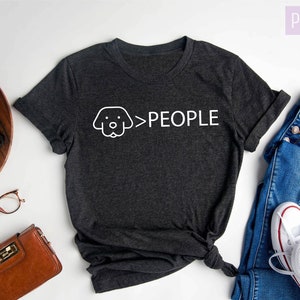 May include: Dark gray heather t-shirt with a white graphic of a dog's head and the text "Dogs > People". The shirt is laid flat on a white surface with a pair of jeans, a wallet, a plant, and a camera.