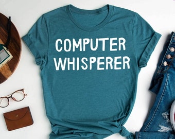 May include: A teal blue t-shirt with the text "COMPUTER WHISPERER" in white. The shirt is laid flat on a white surface with a pair of glasses, a brown wallet, and a pair of blue jeans with white sneakers.
