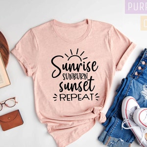 May include: A pink t-shirt with the text "Sunrise Sunburn Sunset Repeat" in black lettering. The shirt is laid out on a white surface with a pair of jeans and white sneakers.