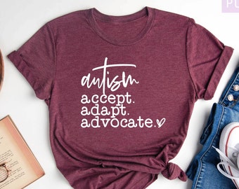 May include: A heather maroon t-shirt with the text "autism accept. adapt. advocate." in white lettering. The shirt is laid flat on a white surface with a book, glasses, and a wooden tray with a candle.