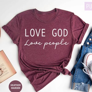 May include: A maroon t-shirt with the text "LOVE GOD Love people" in white lettering. The shirt is laid flat on a white surface with a book, a wooden bowl, and a pair of jeans.
