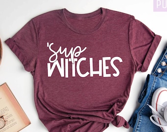 May include: A maroon t-shirt with the text "'Sup Witches" in white script. The shirt is styled with blue jeans and white sneakers. The words "Purple Rush Clothing" and "Heather Maroon" are also visible.