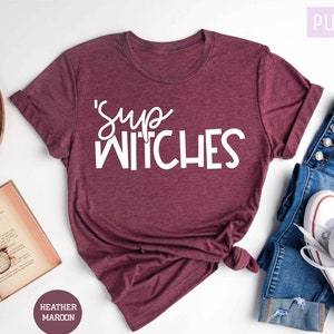Puede incluir: Una camiseta granate con el texto "'Sup Witches" en escritura blanca. La camiseta se combina con vaqueros azules y zapatillas blancas. Tambi&eacute;n se ven las palabras "Purple Rush Clothing" y "Heather Maroon".