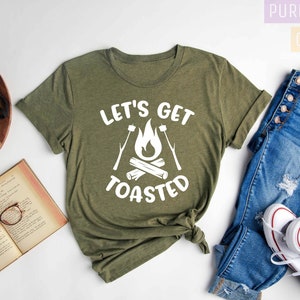 May include: Heather olive green t-shirt with the text "LET'S GET TOASTED" in white. The design features a campfire with marshmallows on sticks. The shirt is paired with blue jeans and white sneakers.