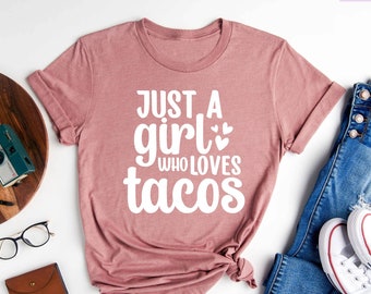 Könnte beinhalten: Ein T-Shirt in Heide-Mauve mit dem Text "Just a girl who loves tacos" in Weiß. Das Shirt liegt flach auf einer weißen Oberfläche mit einer Jeans, einem Paar weißen Sneakers, einem Holzbrett, einer Kamera, einer Brille und einer braunen Ledergeldbörse.