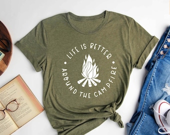 May include: A green t-shirt with a white graphic that says "Life is better around the campfire" with a campfire illustration. The shirt is laid out on a white surface with a book, a pair of glasses, a camera, a plant, and a pair of blue jeans with white trainers.