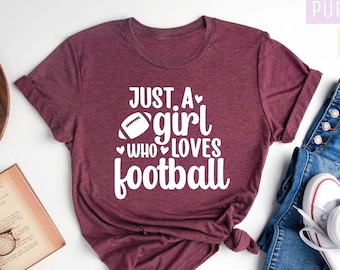 May include: A maroon t-shirt with the text "Just a girl who loves football" in white. The shirt is laid out on a white surface with a book, glasses, and a wooden tray with a candle.