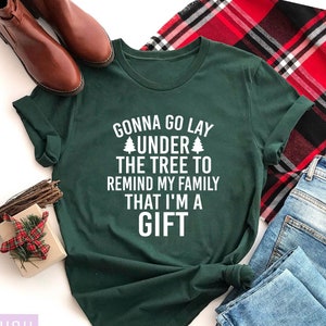 May include: A dark green t-shirt with the text "GONNA GO LAY UNDER THE TREE TO REMIND MY FAMILY THAT I'M A GIFT" in white. The shirt is paired with blue jeans, a red and black plaid blanket, and brown boots. A small gift box is also present.