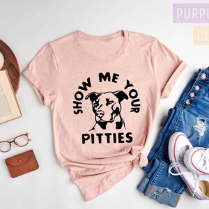 May include: A pink t-shirt with a black graphic that says "Show Me Your Pitties" and features a black silhouette of a Pit Bull dog.