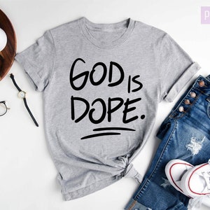 May include: Gray t-shirt with the text "GOD IS DOPE." printed in black. The shirt is shown on a white background with a pair of jeans and white sneakers.
