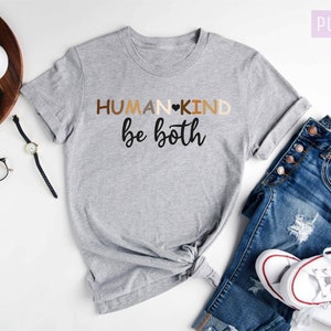 May include: A gray t-shirt with the text "HUMANKIND be both" in black and brown lettering. The shirt is laid out on a white surface with a pair of jeans, a watch, and a camera.