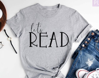May include: A gray t-shirt with the text "let's READ" in black lettering. The shirt is laid out on a white surface with a pair of jeans, a watch, and a camera.
