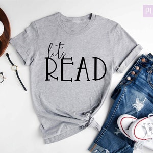 May include: A gray t-shirt with the text "let's READ" in black lettering. The shirt is laid out on a white surface with a pair of jeans, a watch, and a camera.