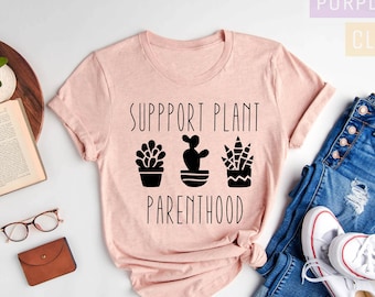May include: A pink t-shirt with the text "Support Plant Parenthood" and three black silhouette images of potted plants. The shirt is laid out on a white surface with a pair of glasses, a book, a small potted plant, a brown wallet, and a pair of blue jeans and white trainers.