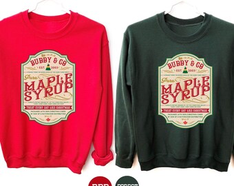 May include: Two crewneck sweatshirts, one red and one forest green, each featuring a vintage-style label design. The label reads "Bubby & Co. Pure Maple Syrup" with text below. The sweatshirts are displayed on white hangers.