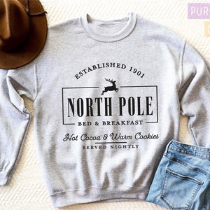 May include: A light gray sweatshirt with the text "NORTH POLE BED & BREAKFAST" in black, along with the words "Hot Cocoa & Warm Cookies Served Nightly." A brown hat and blue jeans are also in the image.