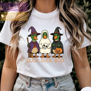 May include: White t-shirt featuring a Halloween-themed graphic. The design includes three cartoon ducks dressed as a witch, a ghost, and a witch with a pumpkin. The text "DUCK, DUCK, BOO" is printed below the ducks.