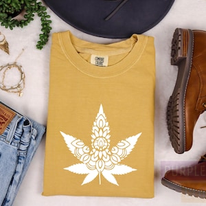 May include: A mustard yellow t-shirt with a white mandala-style marijuana leaf design. The shirt is laid flat on a white surface with other items around it, including a pair of brown leather boots, a pair of jeans, a hat, and jewelry.
