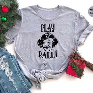 May include: Athletic heather gray t-shirt with the phrase "PLAY BALL!" and a black and white graphic of a woman's face. The shirt is displayed with blue jeans and holiday decorations.