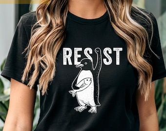 Op de afbeelding: Zwart T-shirt met een witte afbeelding van een pinguïn die een vis vasthoudt en het woord "RESIST".