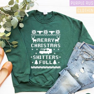 May include: A dark green sweatshirt with a white graphic that reads "MERRY CHRISTMAS *SHITTERS* FULL" with reindeer, snowflakes, a camper, and Christmas trees. The sweatshirt is paired with blue jeans and brown flats.