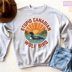 A gray sweatshirt with a graphic design of a loon and a wolf bird, along with the text "STUPID CANADIAN WOLF BIRD" printed on it. The sweatshirt is placed on a bed, and there is a brown hat and a pair of jeans nearby.
