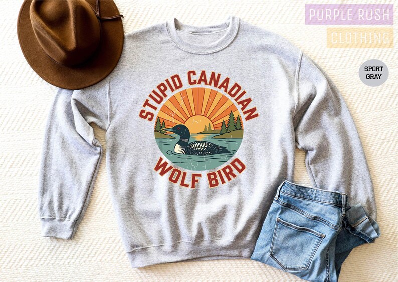 A gray sweatshirt with a graphic design of a loon and a wolf bird, along with the text "STUPID CANADIAN WOLF BIRD" printed on it. The sweatshirt is placed on a bed, and there is a brown hat and a pair of jeans nearby.