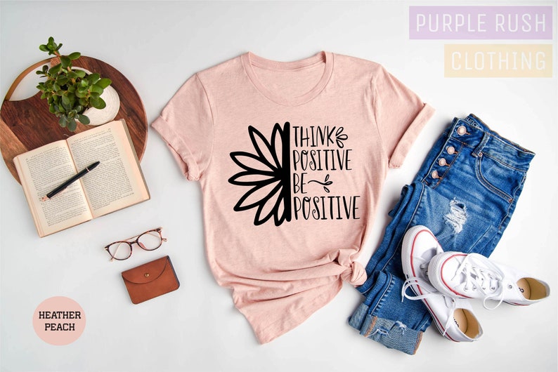 May include: A pink t-shirt with a black graphic of a sunflower and the text "Think Positive Be Positive". The shirt is laid out on a white surface with a pair of jeans, white sneakers, a book, glasses, and a small brown wallet.