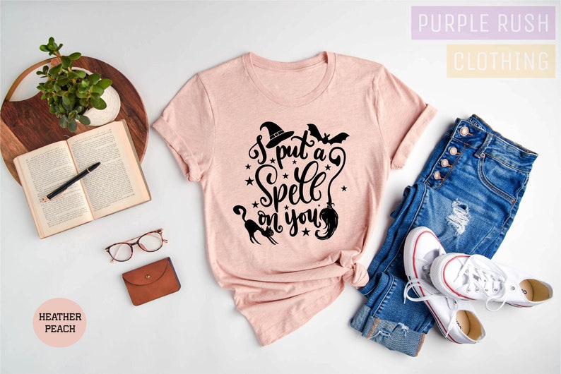 May include: Heather peach-colored t-shirt with the phrase "I put a Spell on you" in black script. The design includes a witch hat, bats, stars, a cat, and a broom. The shirt is paired with blue jeans and white sneakers.