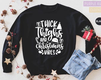 May include: Black sweatshirt with the text "Thick Thighs and Christmas Vibes" in white. The design includes a Christmas tree, a Santa hat, and gift boxes. A pair of blue jeans is folded below the sweatshirt.