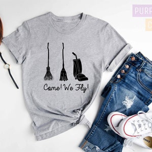 May include: Athletic heather gray t-shirt with a black graphic of a broom, a broom, and a vacuum cleaner. The text "Come! We Fly!" is below the graphics. The shirt is paired with blue jeans and white sneakers.