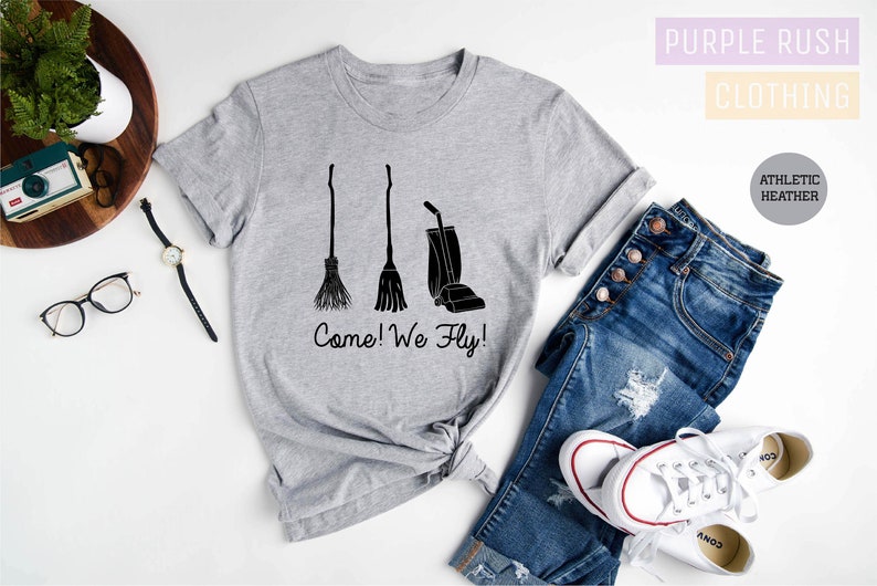 May include: Athletic heather gray t-shirt with a black graphic of a broom, a broom, and a vacuum cleaner. The text "Come! We Fly!" is below the graphics. The shirt is paired with blue jeans and white sneakers.