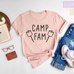 May include: A pink heather peach t-shirt with the text "Camp Fam" and two black marshmallow graphic illustrations. The shirt is laid out on a white surface with a pair of blue jeans and white sneakers.