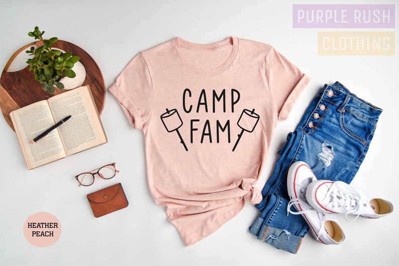 May include: A pink heather peach t-shirt with the text "Camp Fam" and two black marshmallow graphic illustrations. The shirt is laid out on a white surface with a pair of blue jeans and white sneakers.