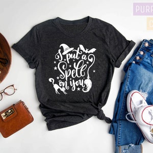 May include: Dark gray heather t-shirt with the text "I put a Spell on you" in white. The design includes a witch hat, bat, stars, a cat, and a broom. The shirt is styled with blue jeans and white sneakers.