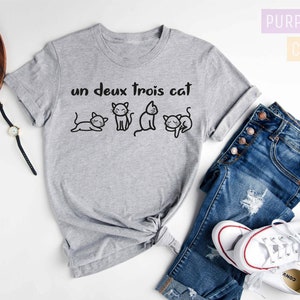 May include: Gray t-shirt with the text "un deux trois cat" and a row of four cartoon cats. The shirt is laid out on a white surface with a pair of jeans, a watch, and a camera.