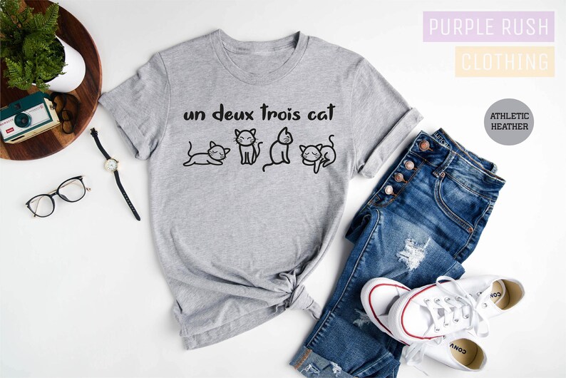 May include: Gray t-shirt with the text "un deux trois cat" and a row of four cartoon cats. The shirt is laid out on a white surface with a pair of jeans, a watch, and a camera.