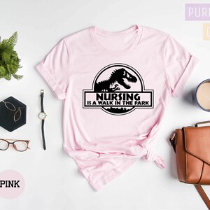 May include: Pink t-shirt with a black graphic of a dinosaur and the text "Nursing is a walk in the park".