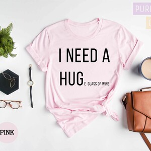 Puede incluir: Camiseta rosa con el texto "Necesito un abrazo y una copa de vino" impreso en negro.