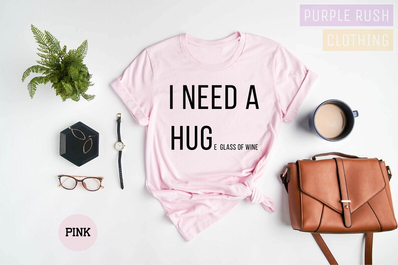 Puede incluir: Camiseta rosa con el texto "Necesito un abrazo y una copa de vino" impreso en negro.