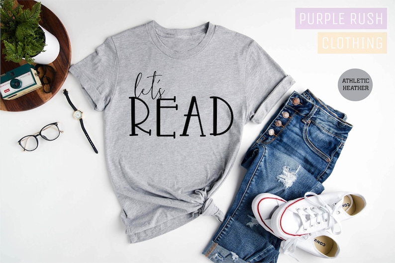 May include: A grey t-shirt with the text "let's READ" in black lettering. The shirt is laid out on a white surface with a pair of jeans, a watch, and a camera.