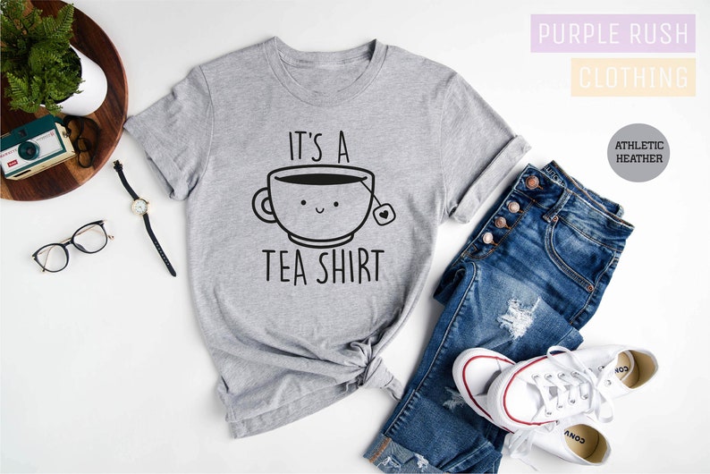May include: A gray t-shirt with the text "It's a Tea Shirt" and a cartoon illustration of a teacup with a smiling face.