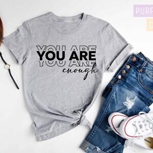 Może przedstawiać: Szary T-shirt z nadrukiem "YOU ARE YOU ARE enough" w kolorze czarnym. Koszulka leży na białym tle z dżinsami i białymi butami sportowymi.