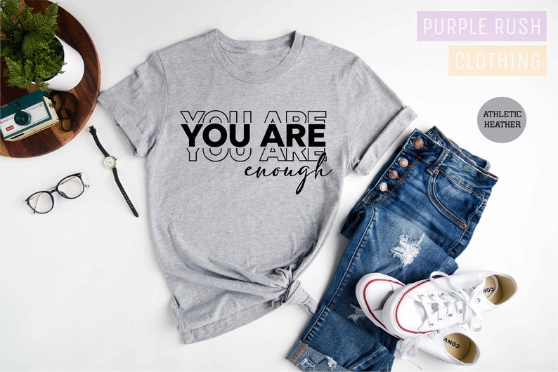 Może przedstawiać: Szary T-shirt z nadrukiem "YOU ARE YOU ARE enough" w kolorze czarnym. Koszulka leży na białym tle z dżinsami i białymi butami sportowymi.