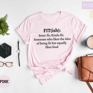 May include: Pink t-shirt with the text "FIT(ish): Semi-fit; Kinda fit; Someone who likes the idea of being fit but equally likes food." The shirt is displayed with accessories including a watch, glasses, and a purse.