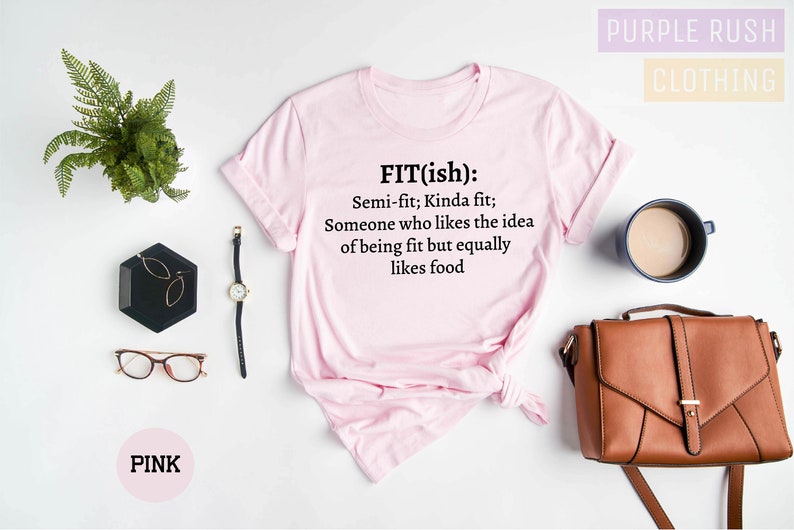 May include: Pink t-shirt with the text "FIT(ish): Semi-fit; Kinda fit; Someone who likes the idea of being fit but equally likes food." The shirt is displayed with accessories including a watch, glasses, and a purse.