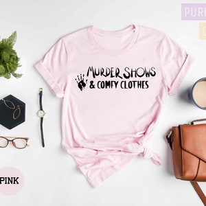 May include: Pink t-shirt with the text "Murder Shows & Comfy Clothes" in black lettering. The shirt is laid flat on a white surface with a black tray, a watch, glasses, a brown purse, and a cup of coffee.