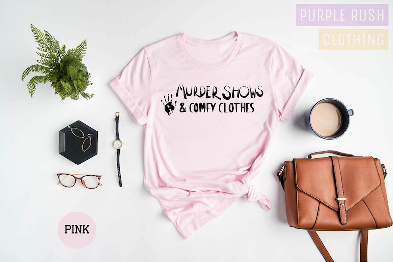 May include: Pink t-shirt with the text "Murder Shows & Comfy Clothes" in black lettering. The shirt is laid flat on a white surface with a black tray, a watch, glasses, a brown purse, and a cup of coffee.