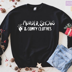 May include: Black sweatshirt with the text "MURDER SHOWS & COMFY CLOTHES" in white. A white handprint graphic is to the left of the text. The sweatshirt is paired with blue jeans. Decorative pinecones, stars, and a small gift box are in the background.