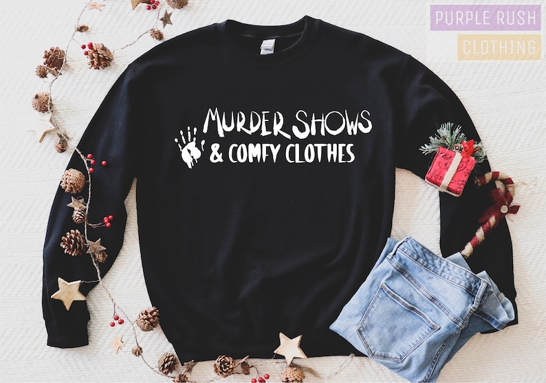May include: Black sweatshirt with the text "MURDER SHOWS & COMFY CLOTHES" in white. A white handprint graphic is to the left of the text. The sweatshirt is paired with blue jeans. Decorative pinecones, stars, and a small gift box are in the background.
